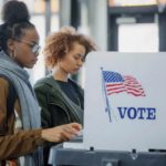 Group of women gathered around a voting machine in a polling station.